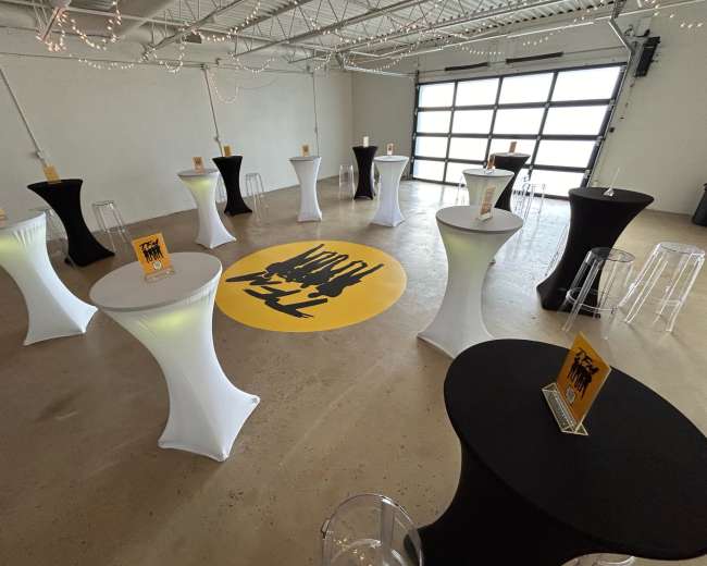 A spacious indoor venue with a polished concrete floor and multiple tall tables adorned with table covers in white and black, along with decorative centerpieces.