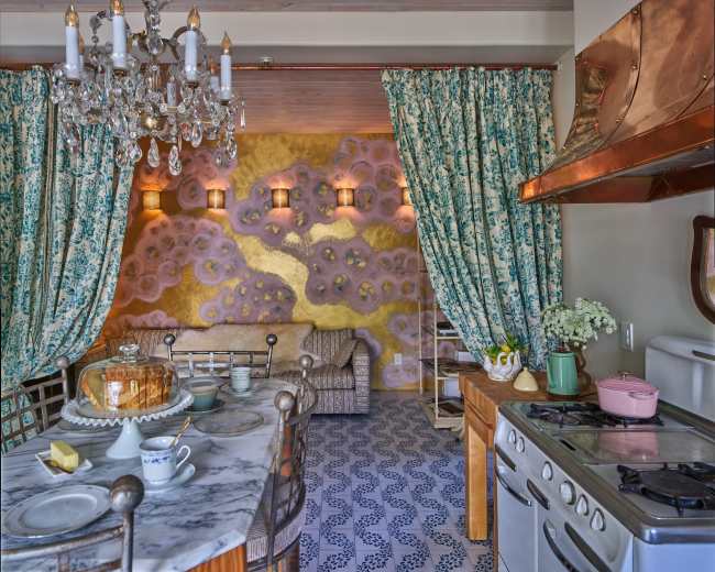 The image shows a stylish kitchen and dining area featuring a marble table, ornate chandelier, and vibrant wall decorations with patterned curtains.