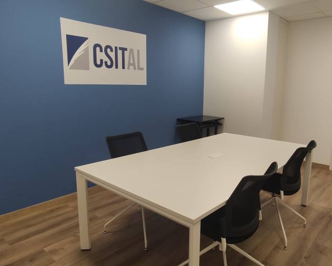 A bright, modern meeting room features a white table with four chairs, a blue wall displaying the "CSITAL" logo, and a door leading to another area.