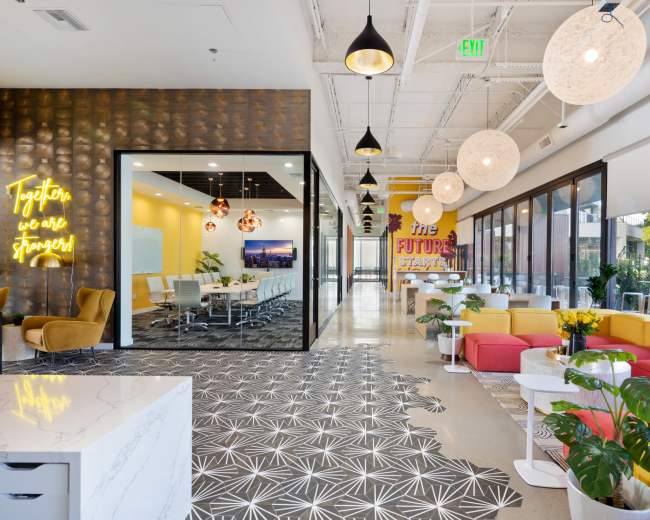The image shows a brightly lit modern office interior featuring a mix of colorful seating, a large meeting room with glass walls, and vibrant wall decor.