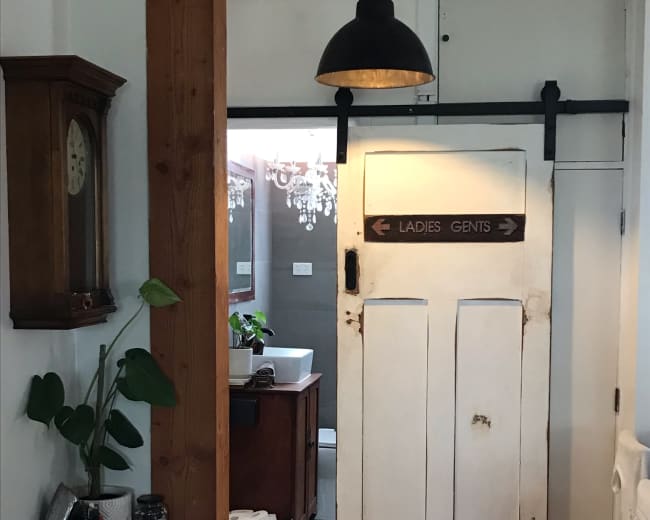 The image shows a hallway leading to a bathroom, featuring a sliding door marked "LADIES" and "GENTS" and elements like a vintage clock and potted plants.