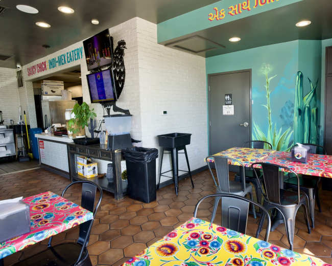 The image shows a brightly colored restaurant interior with patterned tables, a serving area with a menu display, and a mural of plants on the wall.