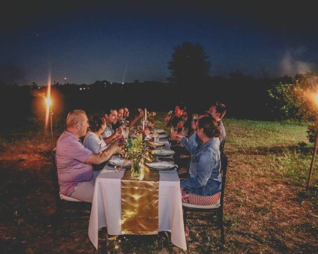 A group of people sits around a long table set for dinner in an outdoor setting at twilight, with tiki torches providing light and a floral centerpiece on the table.