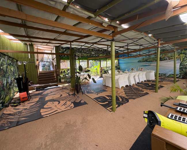 The image shows an indoor event space decorated with tropical murals, featuring tables set for a gathering and a staircase in the background.