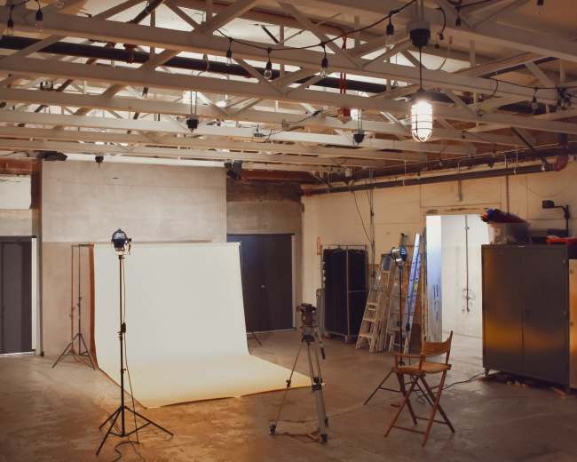 A photography studio features a light-colored backdrop, camera equipment, a director's chair, and exposed beams overhead.