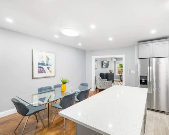 A modern kitchen features a central island with a light countertop, a dining table with chairs, and a stainless steel refrigerator, with a glimpse of a living area in the background.