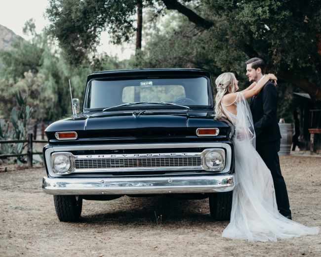 A bride in a flowing gown embraces a groom in a suit in front of a classic black Chevrolet truck.