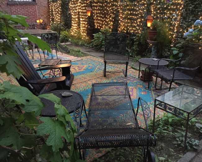 A cozy outdoor seating area features black metal furniture arranged on a colorful patterned rug surrounded by string lights and lush greenery.