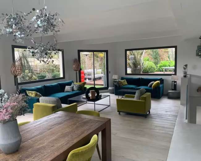 The image depicts a spacious living room with modern furniture, large windows, and views of a garden.