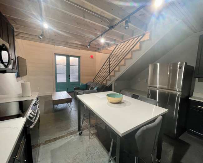 The image shows a modern kitchen with gray cabinetry, a large island with a bowl on it, and a staircase leading to an upper level.