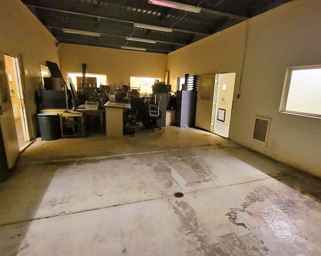 The image shows a dimly lit, empty room with concrete flooring and two glass-paneled doors leading to adjacent areas filled with various equipment and furniture.