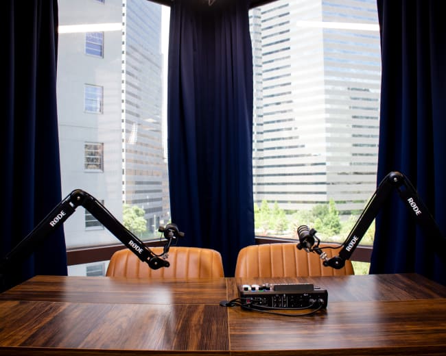 A wooden table with two microphone arms and audio equipment is positioned in front of large windows overlooking city skyscrapers.
