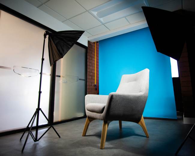 The image features a gray armchair positioned in front of a bright blue backdrop, with two softbox studio lights on either side.