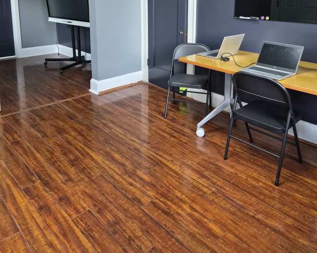 The image shows a room with wooden flooring featuring a desk with two laptops, a large television on a stand, and a door leading to another space.