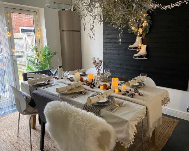 A dining table is set with elegant tableware and decorations, featuring flickering candles, pinecones, and hanging ice skates above.