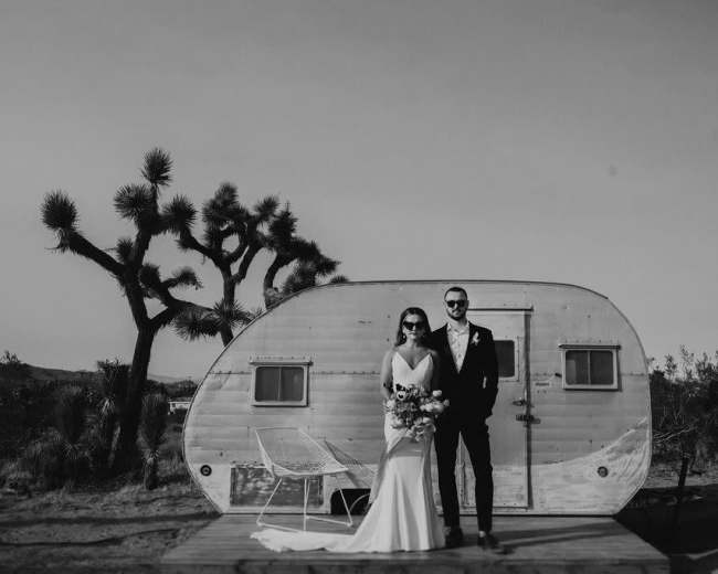 A couple stands in front of a vintage trailer in a desert landscape with Joshua trees.