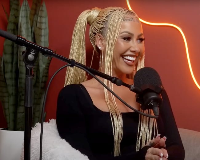 A woman with braided hair smiles while sitting in front of a microphone in a studio with a red wall and a neon light.