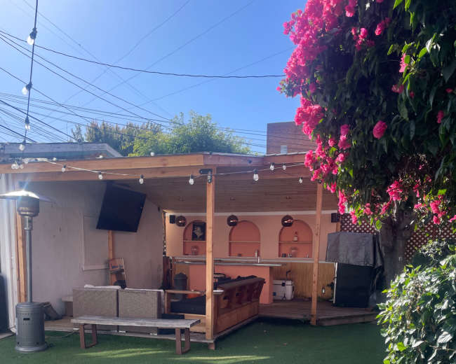 The image shows a backyard area featuring a wooden patio with a couch, a bar setup, and artificial turf, surrounded by greenery and string lights.