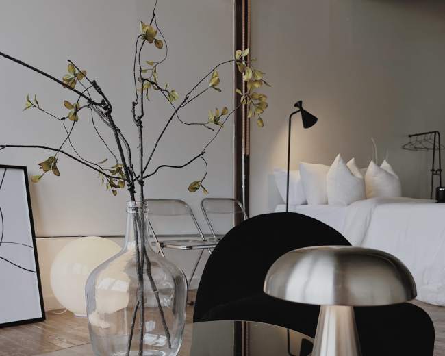 A glass vase with branches sits on a black table next to a metallic mushroom-shaped lamp in a minimalist room.