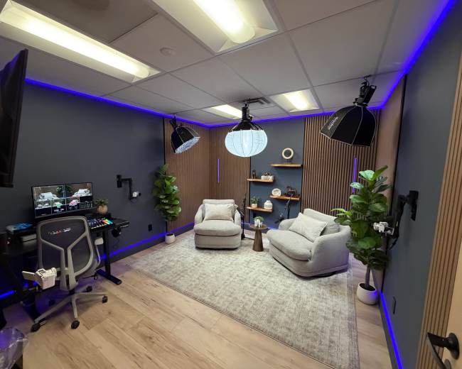The image shows a modern studio room with two grey chairs, a light rug, plants in the corners, and professional lighting setup along with a desk that has computer equipment.