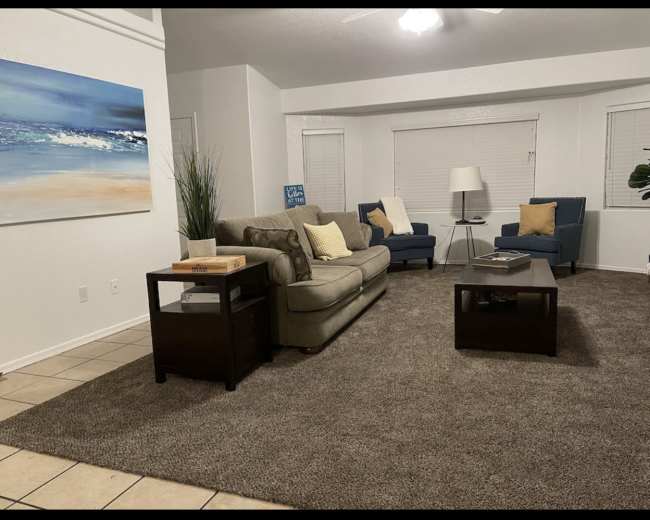 A living room with a beige sofa, two blue armchairs, a dark wooden coffee table, and a large painting of a seascape on the wall.