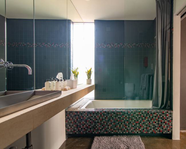 The image shows a modern bathroom with a sunken tub surrounded by mosaic tiles, a sleek sink with toiletries, and a large mirror reflecting a window.