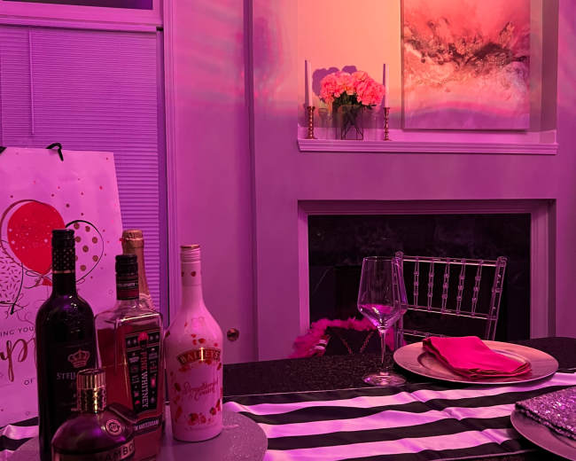 A decorative table set with various liquor bottles, a wine glass, and dinnerware against a softly lit backdrop featuring a framed artwork and flowers.