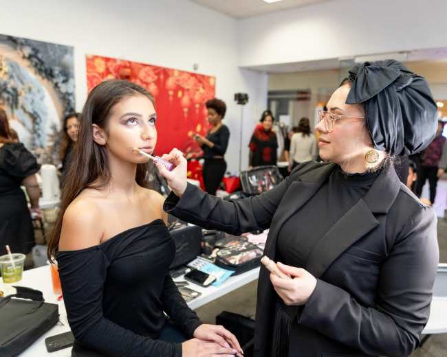 A makeup artist applies makeup to a model in a professional setting with other individuals in the background.