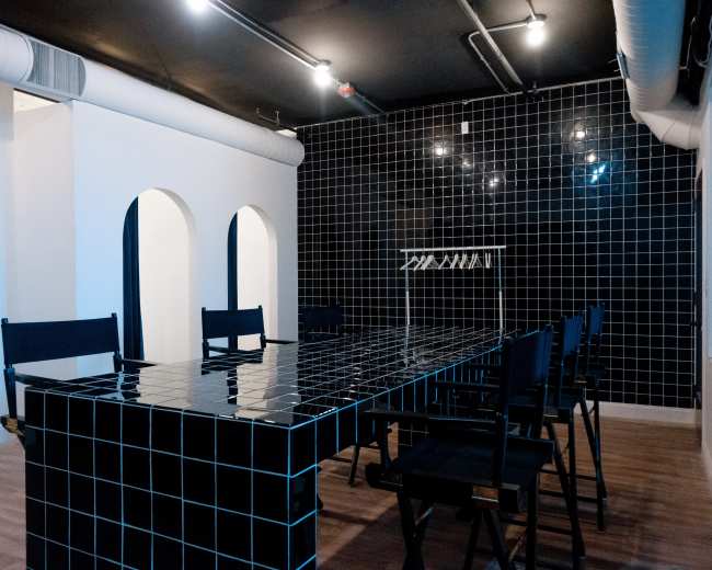 A large, black-tiled table surrounded by black chairs is situated in a room with a contrasting white wall and arched openings, illuminated by overhead lights.