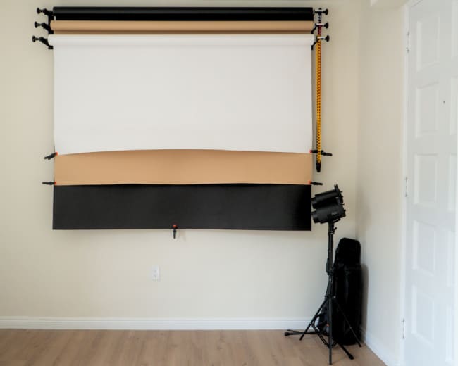 The image shows a blank wall with a photo backdrop setup consisting of three rolls: white, tan, and black, along with a light stand positioned to the side.