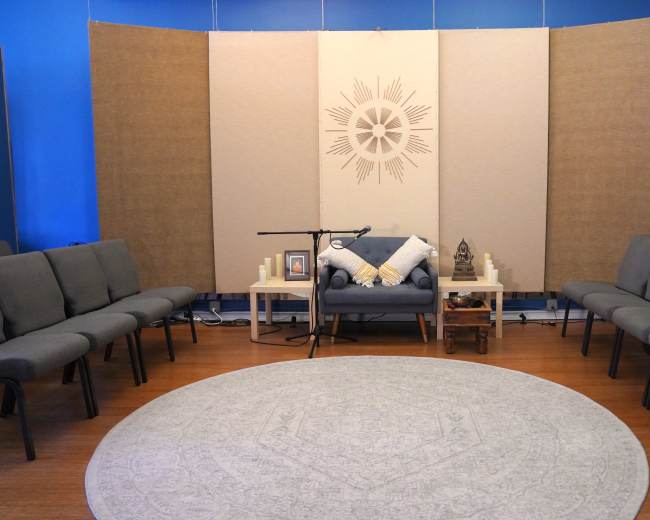 The image shows a serene meditation room with gray seating arranged in a circular formation around a large, circular rug.