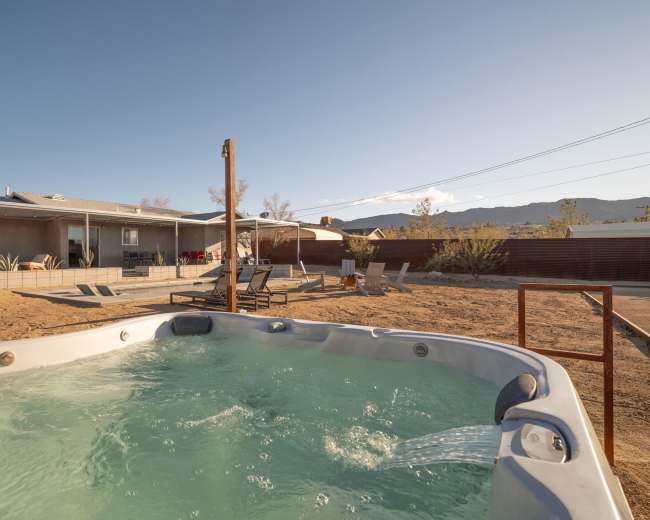 A hot tub with bubbling water is situated in an outdoor area featuring lounge chairs and a house in the background.