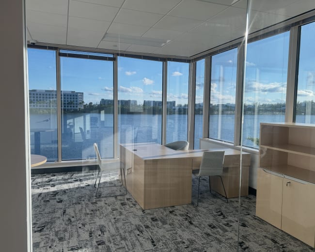 The image shows a modern office space with large windows overlooking a body of water and a view of nearby buildings.