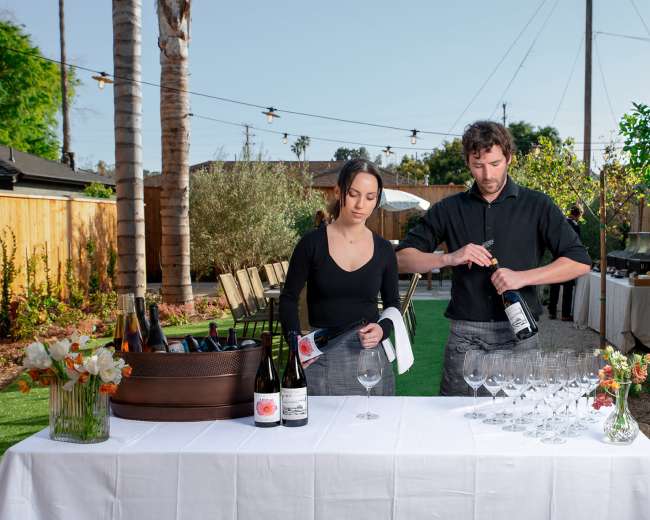 A woman and a man are preparing glasses and pouring wine at an outdoor event with a garden setup.