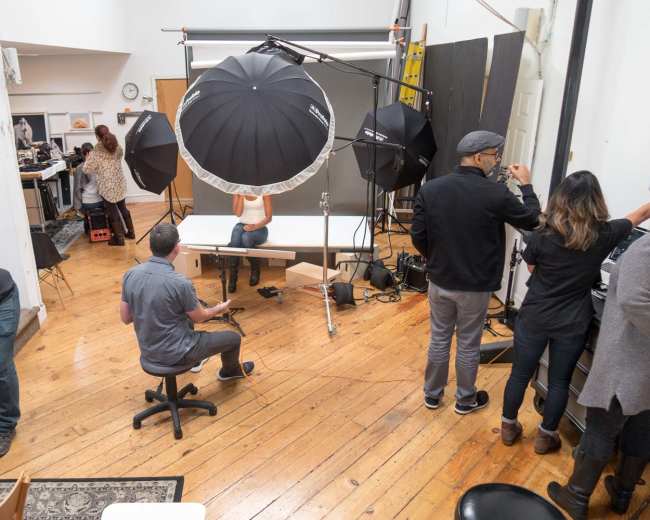 A photography studio is set up with multiple lighting equipment and a model posing on a table while several crew members assist nearby.