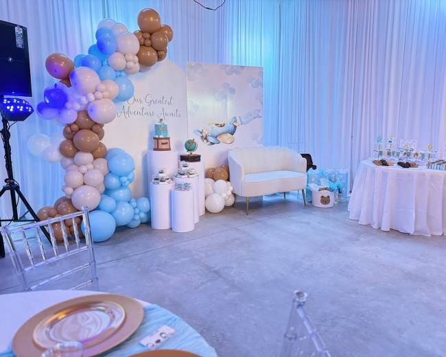 The image shows a decorated event space featuring a balloon arch in blue and brown, a white couch, a dessert table with treats, and a backdrop with a cloud and balloon design.