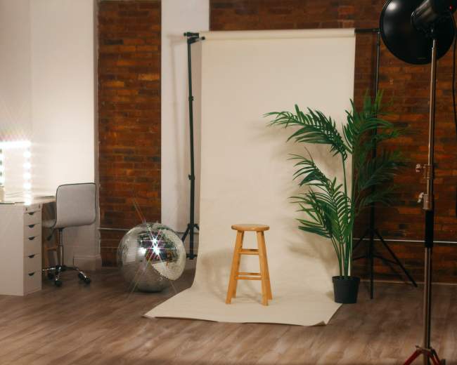 A photography studio with a wooden stool in front of a backdrop, a plant in a pot, a disco ball, and a makeup station with lights.