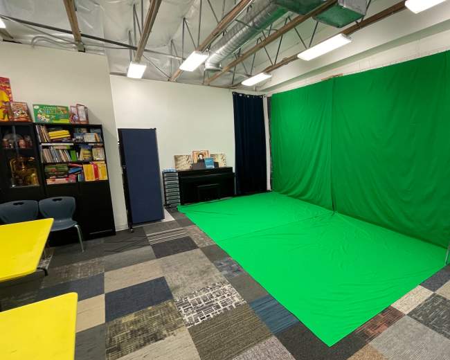A room with a green screen setup against one wall, surrounded by colorful flooring and furniture, including a piano and storage units.