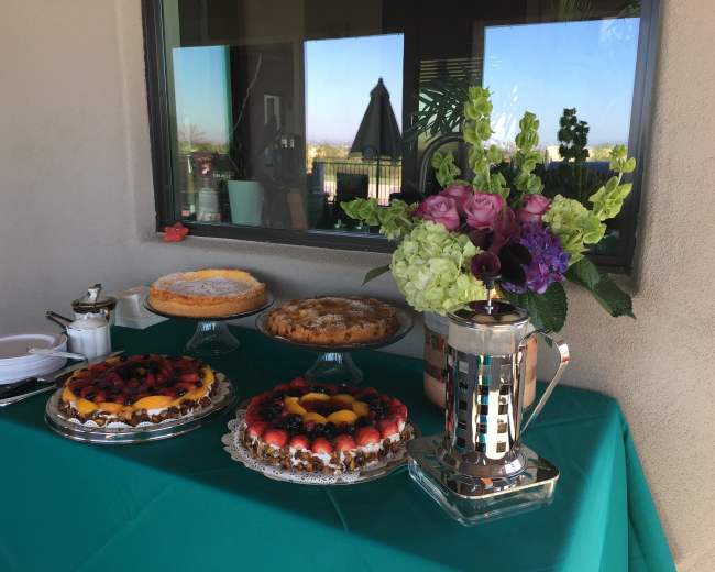 A table is set with various cakes topped with fruit and a floral arrangement next to a coffee press.