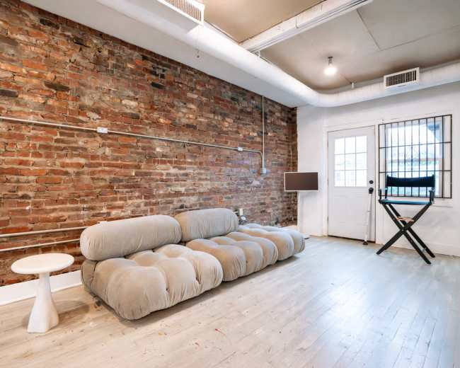 The image shows a spacious room with exposed brick walls, a light-colored wooden floor, a gray sofa, a small white side table, and a door leading outside.