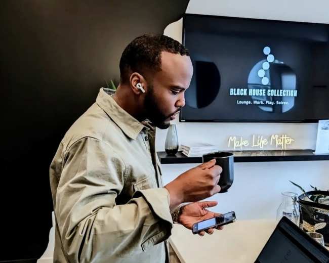 A man is standing in an office environment, holding a mug in one hand while using a smartphone with the other, next to a laptop on a desk.