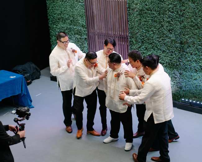 A group of five men in traditional barong shirts celebrate together in front of a green hedge backdrop.