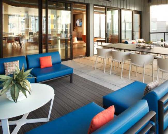 The image shows a modern outdoor lounge area with blue seating, a round table, and a dining table set for several people, surrounded by large glass doors.