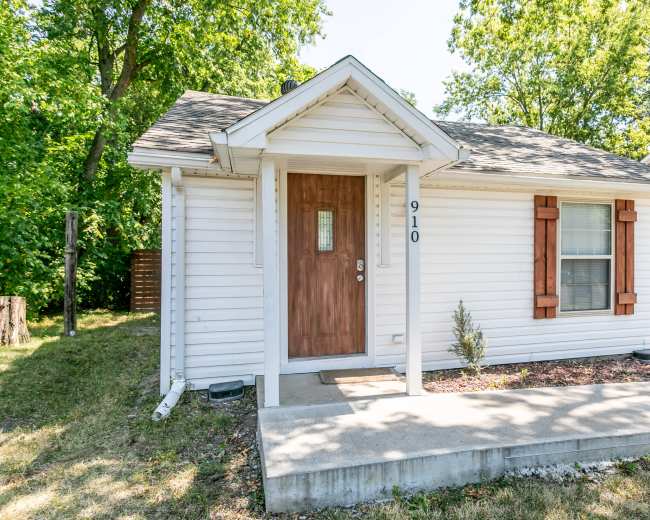 A small, single-story house with a front porch, wooden door, and shutters on the windows, surrounded by green trees and grass.