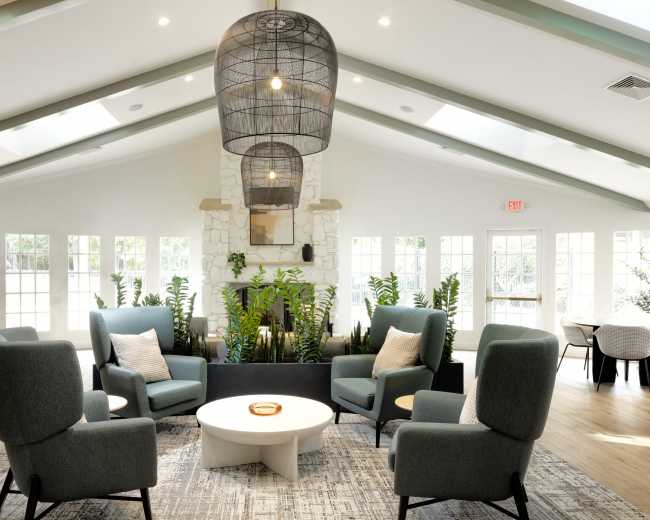 The image shows a spacious living area with four armchairs arranged around a circular coffee table, featuring large windows and indoor plants against a light-colored wall.