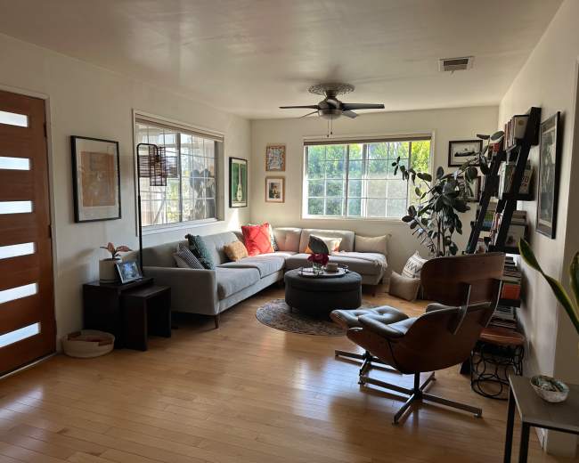 The image shows a living room with a gray sofa, a round coffee table, a wooden chair, and large windows allowing natural light to enter.