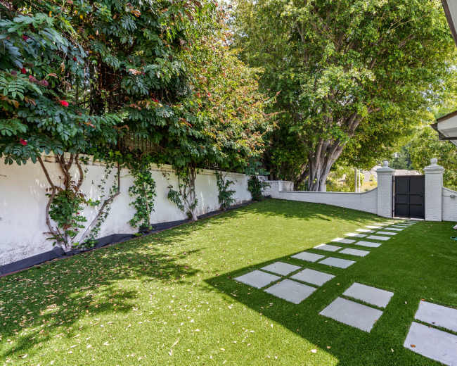 The image shows a well-maintained backyard with artificial grass, a stone pathway, and a wall covered in greenery.