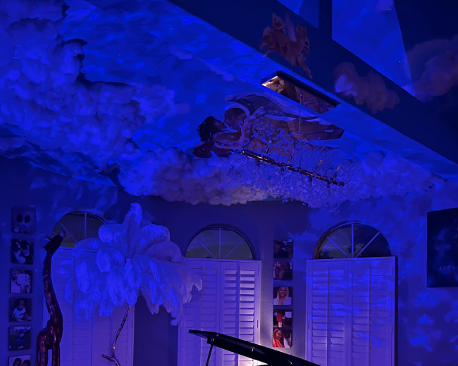 A black grand piano is positioned in a room adorned with cloud-like decorations and blue lighting, surrounded by a large sofa and artistic elements.