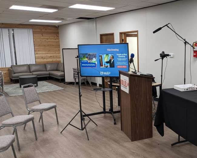 The image shows a meeting room set up with a presentation screen on a stand, a podium with a microphone, and chairs arranged for an audience.