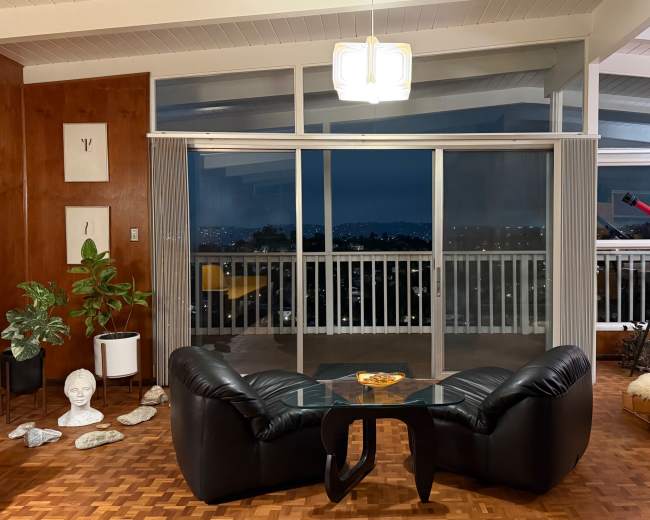 A modern living room features a glass coffee table and black leather armchairs, with a telescope positioned near large sliding glass doors overlooking a city view at dusk.